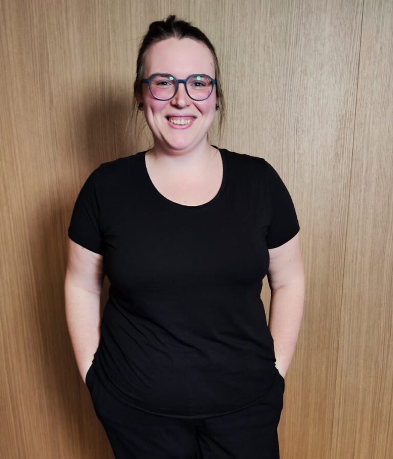 Portrait von Nicci, Projektmanagerin bei Ipsom, mit Brille und schwarzem Shirt, lächelnd vor holzfarbenen Hintergrund