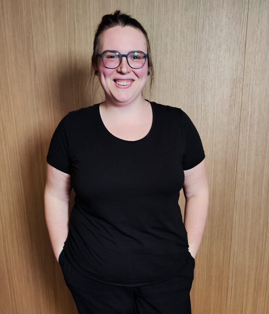 Portrait von Nicci, Projektmanagerin bei Ipsom, mit Brille und schwarzem Shirt, lächelnd vor holzfarbenen Hintergrund
